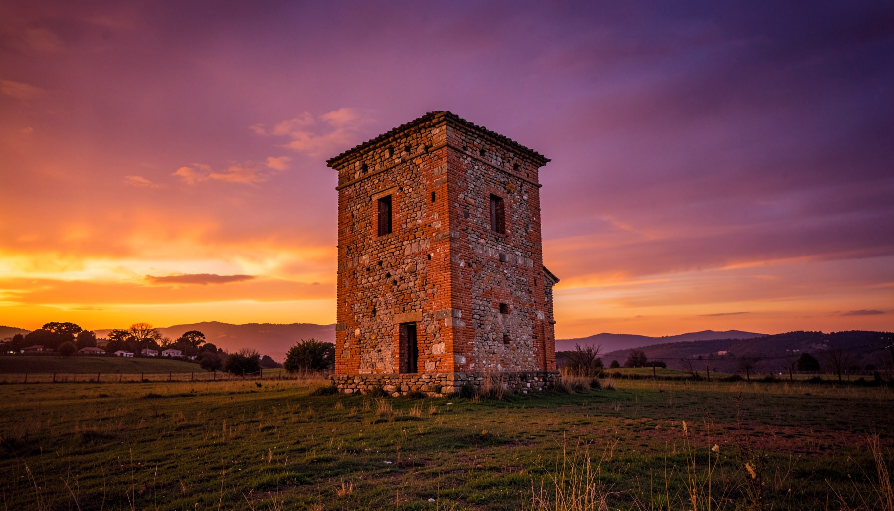 Uma velha torre de pedra e tijolo ergue-se solitária num campo de erva sob um céu dramático de tons púrpura e laranja ao pôr do sol, com colinas distantes no horizonte.
